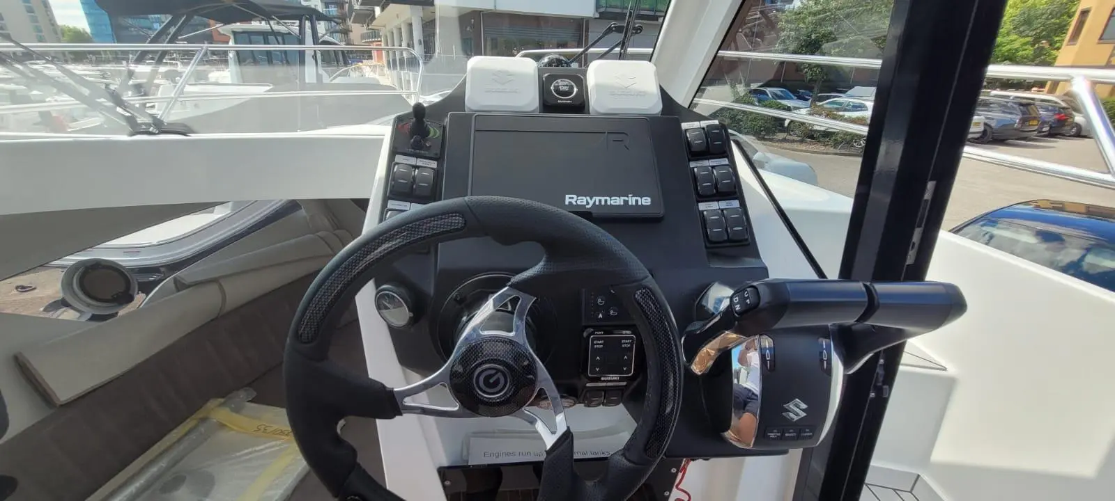 Boat cockpit featuring a Raymarine display, steering wheel, and control levers, with visible logos and adjacent boats.